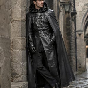 May include: A black leather long coat featuring a hood, a high collar, and a front closure with four horizontal buckled straps. The garment includes long sleeves, matching black leather gloves, and a waist belt with a buckle, worn over black trousers.