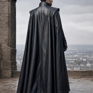 May include: A floor-length black leather cape featuring a high collar, structured shoulder panels, and a smooth finish. The garment drapes down to the ankles, creating a full silhouette with vertical seam detailing along the back and sides.