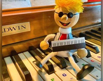 Peut inclure: Une figurine en bois fantaisiste avec des cheveux jaunes, des lunettes rondes et une cravate rouge, assise sur un clavier de piano. La figurine tient un clavier miniature et est positionnée sur les touches du piano. Les touches du piano sont étiquetées avec des notes de musique.