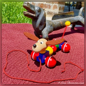 May include: A wooden pull toy dog with red, yellow, blue, and green wheels. The dog has brown ears, a brown nose, and a blue body. The toy is on a red rug with a black cast iron dog in the background.