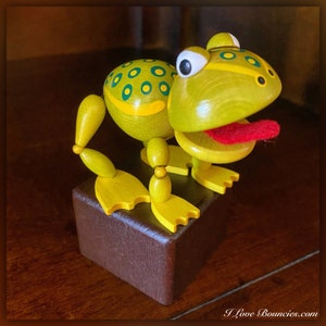 May include: A wooden frog toy with a yellow body, green spots, and a red tongue. The frog is sitting on a brown wooden base.