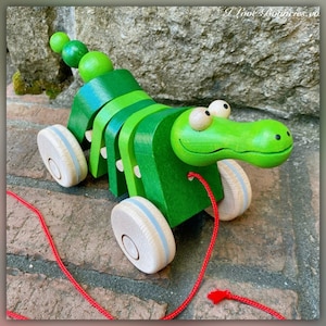 May include: A green wooden pull toy shaped like an alligator with large eyes and a long snout. The toy has four wheels and a red string attached to the back.