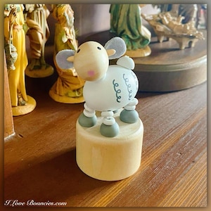May include: A white wooden toy sheep with gray ears and legs. The sheep is standing on a wooden base and has a black curly design on its side. The sheep is a traditional Christmas decoration.