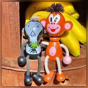 Puede incluir: Dos figuras de mono de madera con extremidades de cuerda y detalles pintados. Una es gris y negra con un mechón de pelo, la otra es naranja y rosa con un mechón de pelo. Están frente a plátanos amarillos. El texto I Love Bouncies.com es visible.