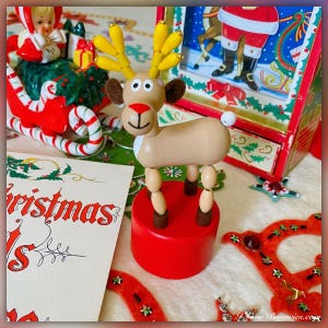 May include: A wooden reindeer toy with a red nose, brown eyes, and yellow antlers. The reindeer is standing on a red base and is surrounded by Christmas decorations.
