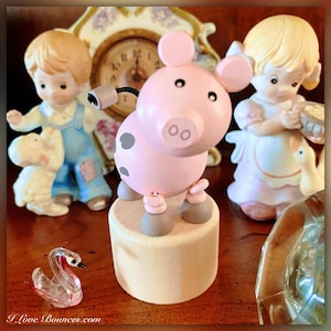 May include: A pink wooden pig with black spots and a gray base. The pig is on a wooden base and has a black tail. The pig is a toy and is in focus. The pig is in front of a clock and two porcelain figurines.