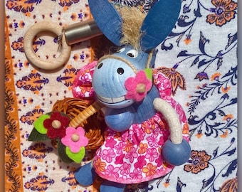 Könnte beinhalten: Eine skurrile, blaue Eselfigur mit großen Ohren, die ein rosa Blumenkleid trägt und einen Weidenkorb mit Blumen hält. Der Esel hat einen lächelnden Gesichtsausdruck und ist mit einem Holzring mit einer Metallfeder verbunden. Der Hintergrund zeigt einen gemusterten Stoff.
