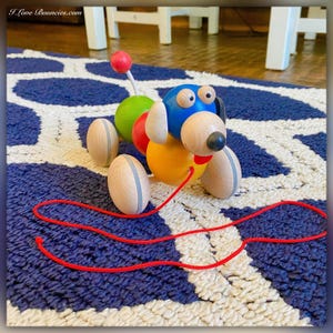 May include: A colorful wooden pull toy shaped like a dog with a red string. The dog has blue, yellow, red, and green painted details. The toy is on a blue and white rug.
