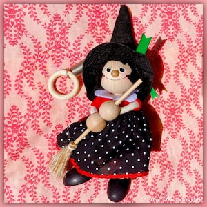 May include: A wooden witch doll with a black hat, red dress, and white polka dots. The doll is holding a wooden broom and a ring. The doll is sitting on a pink and white patterned fabric.