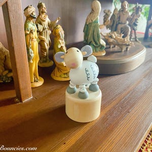 以下が含まれることがあります： 白木製の羊の玩具で、グレーのアクセントと白黒のデザインが施されています。羊は木製の台座の上に置かれており、クリスマスの飾り付けの一部です。