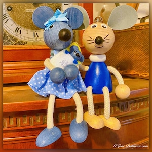 May include: Two wooden mouse figurines. One is blue with a polka dot dress and a bow, holding a small stuffed mouse. The other is natural wood with a blue top. Both have large ears and long legs.