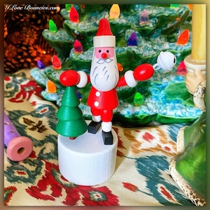 May include: A wooden Santa Claus figurine with red and white clothing, holding a bell in one hand and a small green Christmas tree in the other. The figurine is standing on a white base.