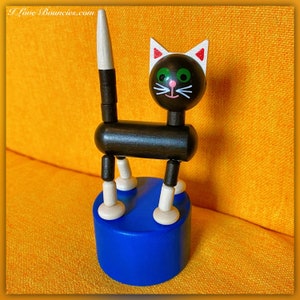 Könnte beinhalten: Ein Holzspielzeug in Form einer Katze mit schwarzem Körper, Kopf und Schwanz sowie weißen und beigefarbenen Akzenten. Die Katze hat grüne Augen, eine rosa Nase und einen weißen Schnurrbart. Sie steht auf einem blauen Sockel. Der Text "I Love Bouncies.com" befindet sich oben links.