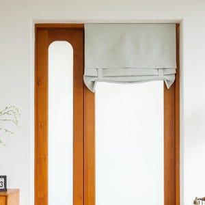 May include: A light gray roman shade hangs in a window with a wooden frame. The shade is pulled up to the top of the window, revealing the white wall behind it.
