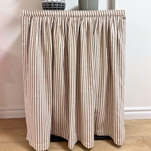 May include: A cabinet with a striped curtain in shades of brown and white. On top, a gray vase with greenery, and a gray candle with the text "SEA SAND". The cabinet is placed against a white wall and a wooden floor.