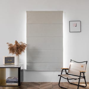 Puede incluir: Una estor romana gris claro con cinco paneles horizontales cuelga de una ventana. Una peque&ntilde;a mesa auxiliar con libros y una radio se encuentra delante de la ventana. Una silla plegable con un asiento de tela beige se encuentra en primer plano.
