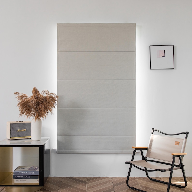 Puede incluir: Una estor romana gris claro con cinco paneles horizontales cuelga de una ventana. Una peque&ntilde;a mesa auxiliar con libros y una radio se encuentra delante de la ventana. Una silla plegable con un asiento de tela beige se encuentra en primer plano.