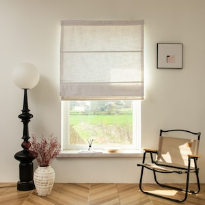 May include: A white linen Roman shade with two folds hangs in a window with a view of a green field. A black floor lamp with a white globe shade and a vase with dried red flowers are on the left side of the window. A beige folding chair is on the right side of the window.