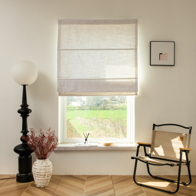 May include: A white linen Roman shade with two folds hangs in a window with a view of a green field. A black floor lamp with a white globe shade and a vase with dried red flowers are on the left side of the window. A beige folding chair is on the right side of the window.