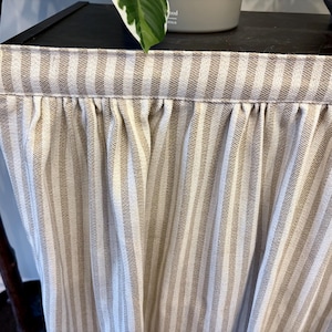 K&ouml;nnte beinhalten: Nahaufnahme eines beige-wei&szlig; gestreiften Stoffes, wahrscheinlich ein Vorhang oder eine Fensterblende. Der Stoff ist oben gerafft, wodurch weiche Falten entstehen. Die vertikalen Streifen sind gleichm&auml;&szlig;ig angeordnet und verleihen dem Textil einen klassischen Touch.