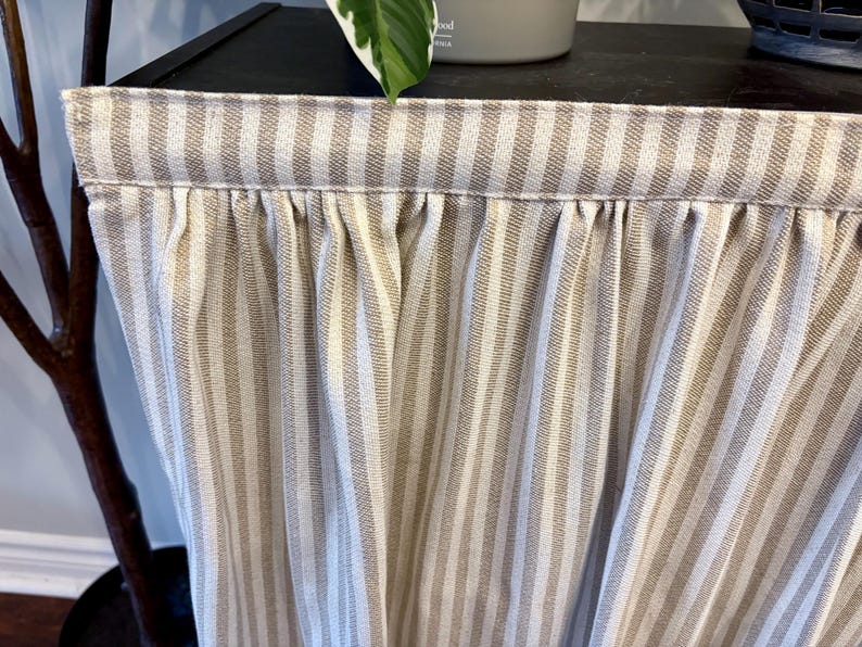 K&ouml;nnte beinhalten: Nahaufnahme eines beige-wei&szlig; gestreiften Stoffes, wahrscheinlich ein Vorhang oder eine Fensterblende. Der Stoff ist oben gerafft, wodurch weiche Falten entstehen. Die vertikalen Streifen sind gleichm&auml;&szlig;ig angeordnet und verleihen dem Textil einen klassischen Touch.