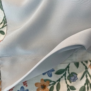 May include: A close-up of a light blue fabric with a subtle texture. The fabric is folded over a white fabric with a floral pattern.