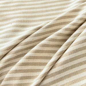 K&ouml;nnte beinhalten: Nahaufnahme eines beige-wei&szlig; gestreiften Stoffes. Der Stoff ist gefaltet, wodurch weiche Schatten entstehen und die Textur des Materials hervorgehoben wird. Die Streifen sind gleichm&auml;&szlig;ig verteilt und verlaufen horizontal &uuml;ber das Bild.