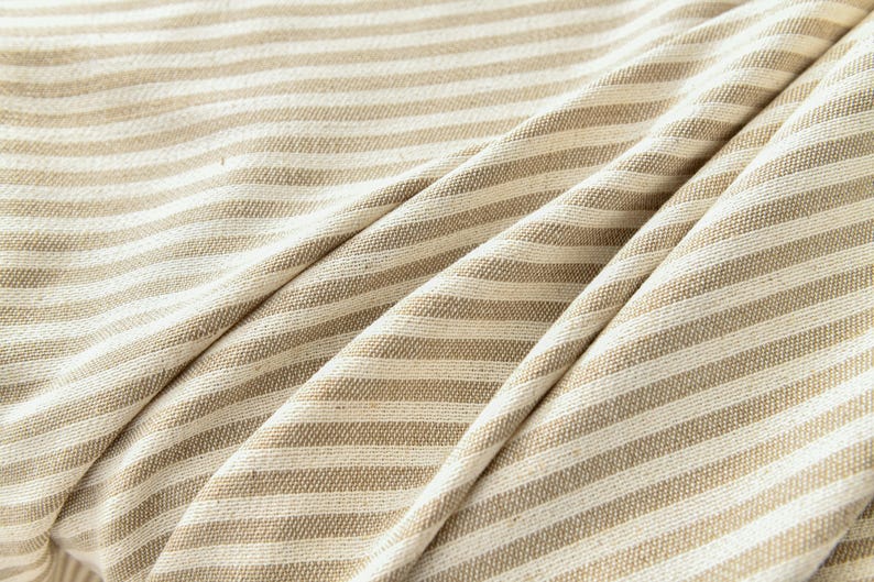 K&ouml;nnte beinhalten: Nahaufnahme eines beige-wei&szlig; gestreiften Stoffes. Der Stoff ist gefaltet, wodurch weiche Schatten entstehen und die Textur des Materials hervorgehoben wird. Die Streifen sind gleichm&auml;&szlig;ig verteilt und verlaufen horizontal &uuml;ber das Bild.