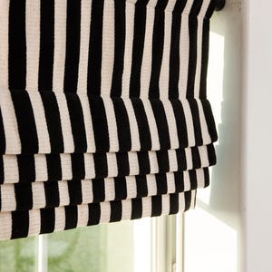 May include: Black and white striped roman shade window covering. The shade is pulled up and the window is open, showing a view of green foliage outside.