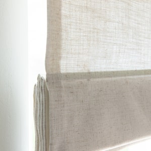 May include: A white linen roman shade with a subtle texture. The shade is partially rolled up, revealing the fabric underneath.