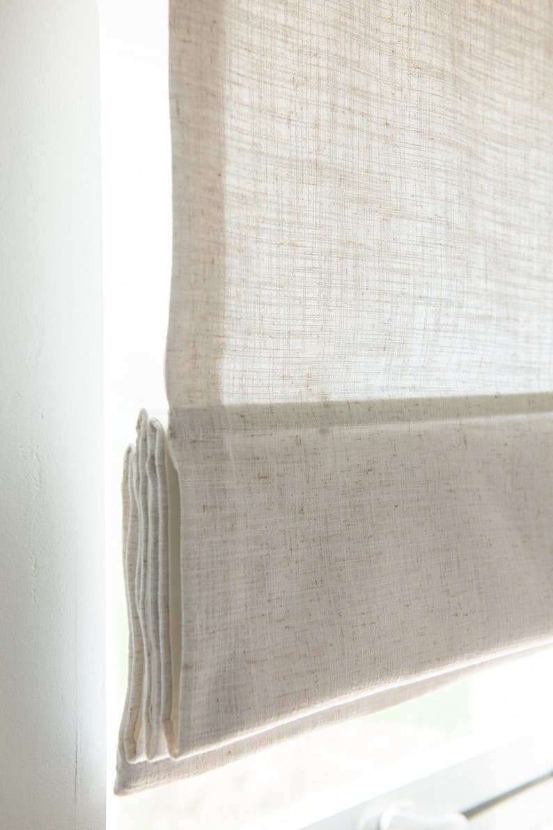 May include: A white linen roman shade with a subtle texture. The shade is partially rolled up, revealing the fabric underneath.