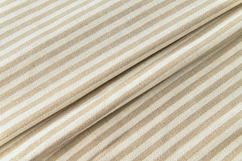 K&ouml;nnte beinhalten: Nahaufnahme eines gefalteten Stoffes mit einem sich wiederholenden Muster aus beige- und cremefarbenen Streifen. Die diagonalen Falten erzeugen Schatten und Tiefe und heben die Textur des Materials hervor. Die Streifen verlaufen leicht schr&auml;g.