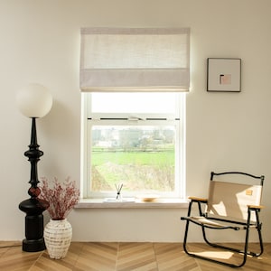 May include: A white linen roman shade with a window view of a green field and trees. A white framed picture hangs on the wall to the right of the window. A black floor lamp with a white globe shade is on the left side of the window. A beige folding chair is on the right side of the window.