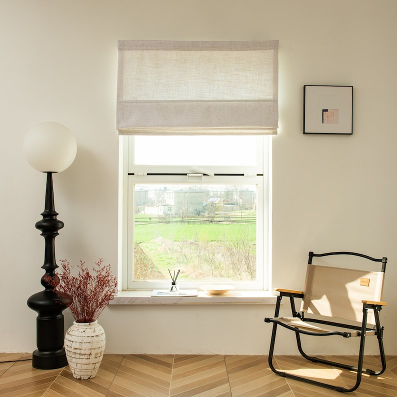 May include: A white linen roman shade with a window view of a green field and trees. A white framed picture hangs on the wall to the right of the window. A black floor lamp with a white globe shade is on the left side of the window. A beige folding chair is on the right side of the window.