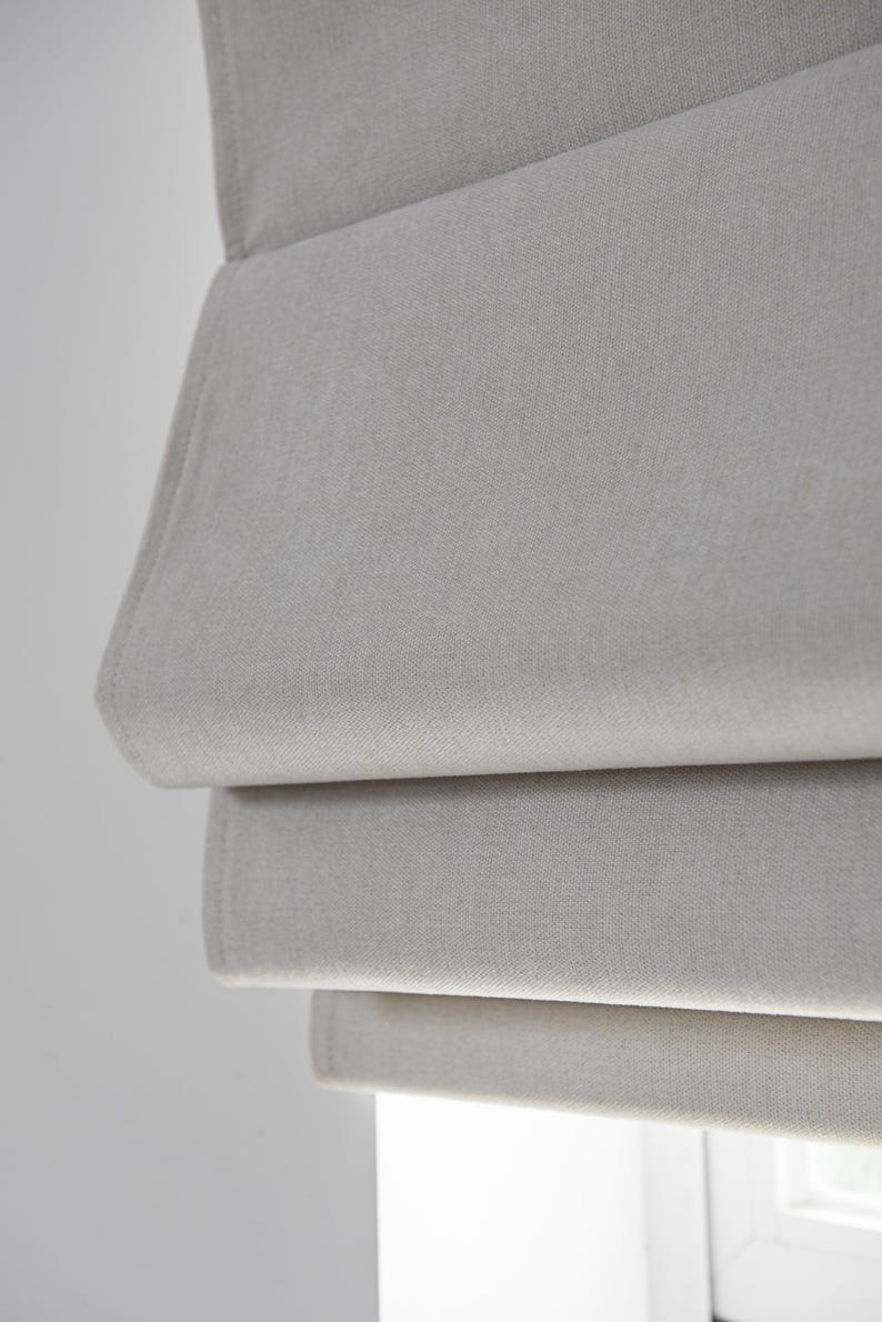 Puede incluir: Primer plano de una estor romano de color gris claro con tejido texturizado. El estor est&aacute; parcialmente subido, mostrando las capas de tejido.