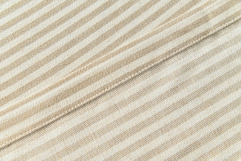 K&ouml;nnte beinhalten: Nahaufnahme eines beige-wei&szlig; gestreiften Stoffes. Der Stoff hat ein diagonales Muster mit abwechselnd beigen und wei&szlig;en Streifen. Der Stoff ist gefaltet und zeigt eine Naht.