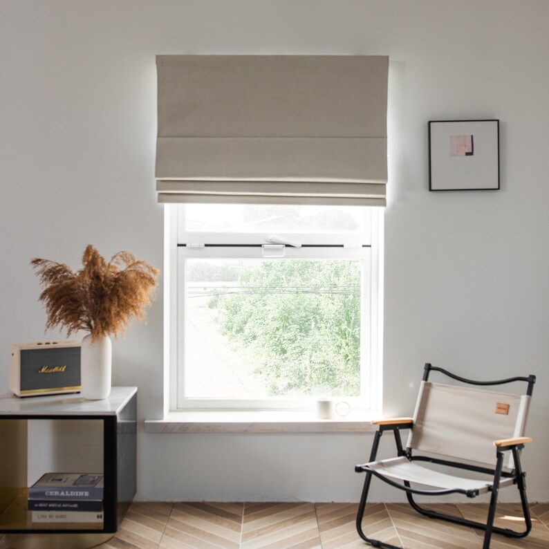 Puede incluir: Una cortina romana beige cuelga en una ventana con vista a &aacute;rboles verdes. Una silla blanca con un marco negro se encuentra en la habitaci&oacute;n. Una mesa peque&ntilde;a con una tapa negra y patas doradas se encuentra junto a la ventana. Un jarr&oacute;n blanco con hierba de la pampa marr&oacute;n seca se encuentra sobre la mesa. Un altavoz negro y dorado se encuentra sobre la mesa.
