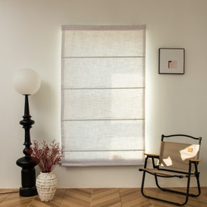 May include: A white linen Roman shade with three horizontal slats hangs in a window. A black floor lamp with a white globe shade sits to the left of the window. A brown folding chair with a beige fabric seat sits to the right of the window. A small framed picture hangs on the wall to the right of the window.
