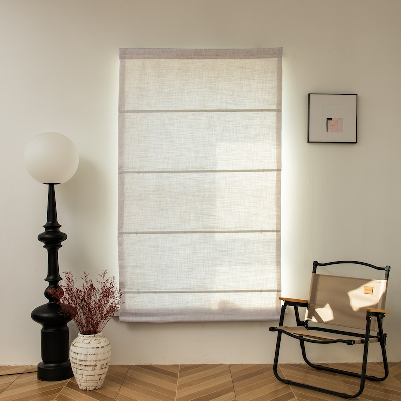May include: A white linen Roman shade with three horizontal slats hangs in a window. A black floor lamp with a white globe shade sits to the left of the window. A brown folding chair with a beige fabric seat sits to the right of the window. A small framed picture hangs on the wall to the right of the window.