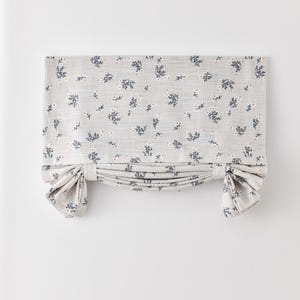 May include: A grey floral roman blind featuring a pattern of small blue flowers and leaves. The blind is tied up on both sides, creating a decorative, gathered appearance. The fabric appears to be linen.