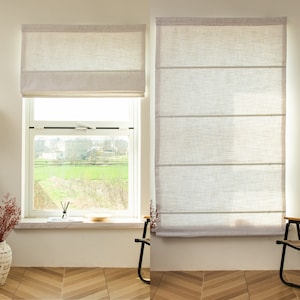 May include: Two white linen roman shades with a light gray trim. The shades are pulled up to reveal a window with a view of a green field and trees.
