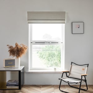 Puede incluir: Una ventana blanca con una cortina romana beige, un cuadro enmarcado en negro en la pared, una mesita auxiliar con un libro y un altavoz, y una silla plegable beige.