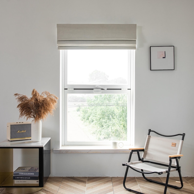 Puede incluir: Una ventana blanca con una cortina romana beige, un cuadro enmarcado en negro en la pared, una mesita auxiliar con un libro y un altavoz, y una silla plegable beige.