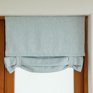 May include: A light blue roman shade window covering with a textured fabric. The shade is pulled up and gathered at the sides.