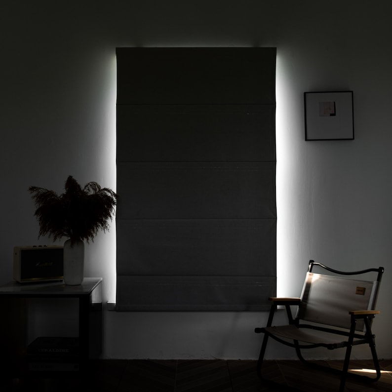 Puede incluir: Una persiana de ventana gris oscuro con una luz blanca que brilla a trav&eacute;s de ella. La persiana est&aacute; hecha de tela y tiene un dise&ntilde;o de listones horizontales. Una silla est&aacute; en primer plano y un jarr&oacute;n con flores secas est&aacute; en una mesa en el fondo.