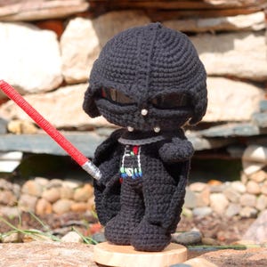 Könnte beinhalten: Handgefertigte Darth Vader-Puppe aus Häkelarbeit, mit schwarzem Helm, Umhang und Körper. Die Puppe hält ein rotes Lichtschwert. Die Figur steht auf einem Holzsockel, mit einem Steinhintergrund.