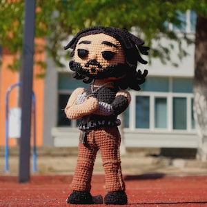 Può includere: Bambola all'uncinetto fatta a mano raffigurante un uomo con dreadlocks neri e barba, vestito con un abito nero e beige con le braccia incrociate. La bambola è in piedi su una superficie rossa, con uno sfondo sfocato di un edificio e alberi.