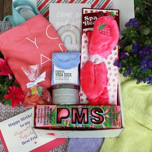 May include: A gift box filled with self-care items. Includes a pink tote bag, a candle, a pair of pink spa headbands, a bag of candy, a pair of yoga socks, and a card that says "Happy Birthday to my Crazy and Truly Amazing Best Friend".