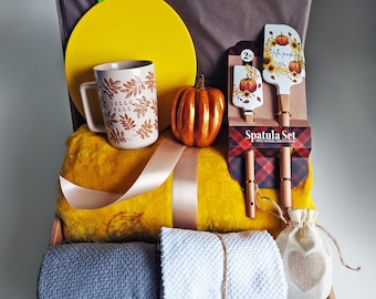 May include: A gift basket filled with fall-themed items. Includes a yellow cutting board, a mug with "Hello Fall" text, a decorative pumpkin, a yellow blanket, a spatula set, and rolled towels. A small bag with a heart is also included.