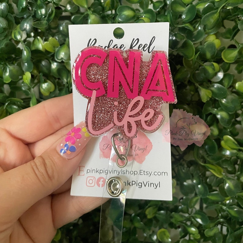 May include: A pink CNA Life badge reel with glitter detailing. The badge reel features the words "CNA Life" in a layered design. The badge reel is attached to a clear plastic holder with a metal clip.